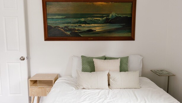 Une chambre à coucher comprenant un lit, des oreillers, une table de chevet, une petite table et un tableau encadré représentant un paysage marin.