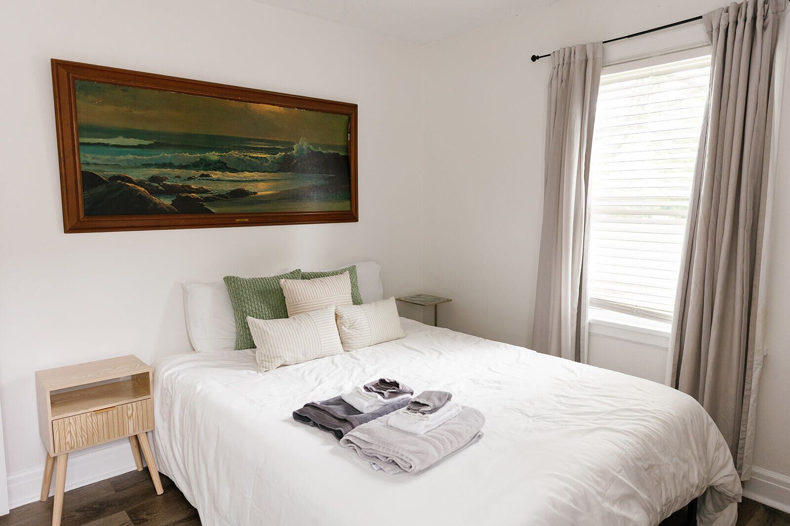 Een net opgemaakt bed met witte lakens en een gevouwen handdoek, een klein nachtkastje en een schilderij van een landschap erboven.