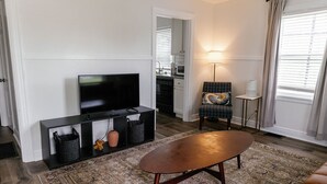 Un salon avec un téléviseur à écran plat posé sur un meuble noir, un tapis à motifs, une table basse en bois, un fauteuil à carreaux et une petite table d’appoint avec une lampe.