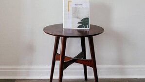 Une table ronde en bois, sur laquelle est posée une boîte blanche portant l’inscription « BIENVENUE ! », est placée contre un mur uni, orné de trois crochets noirs et d’une plante verte.