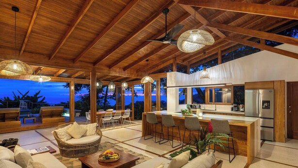A spacious living area with a wooden ceiling, a dining set, and a kitchen area with a refrigerator. - Casa Rica _ Stunning Ocean Views and Private Pool (Cóbano)
