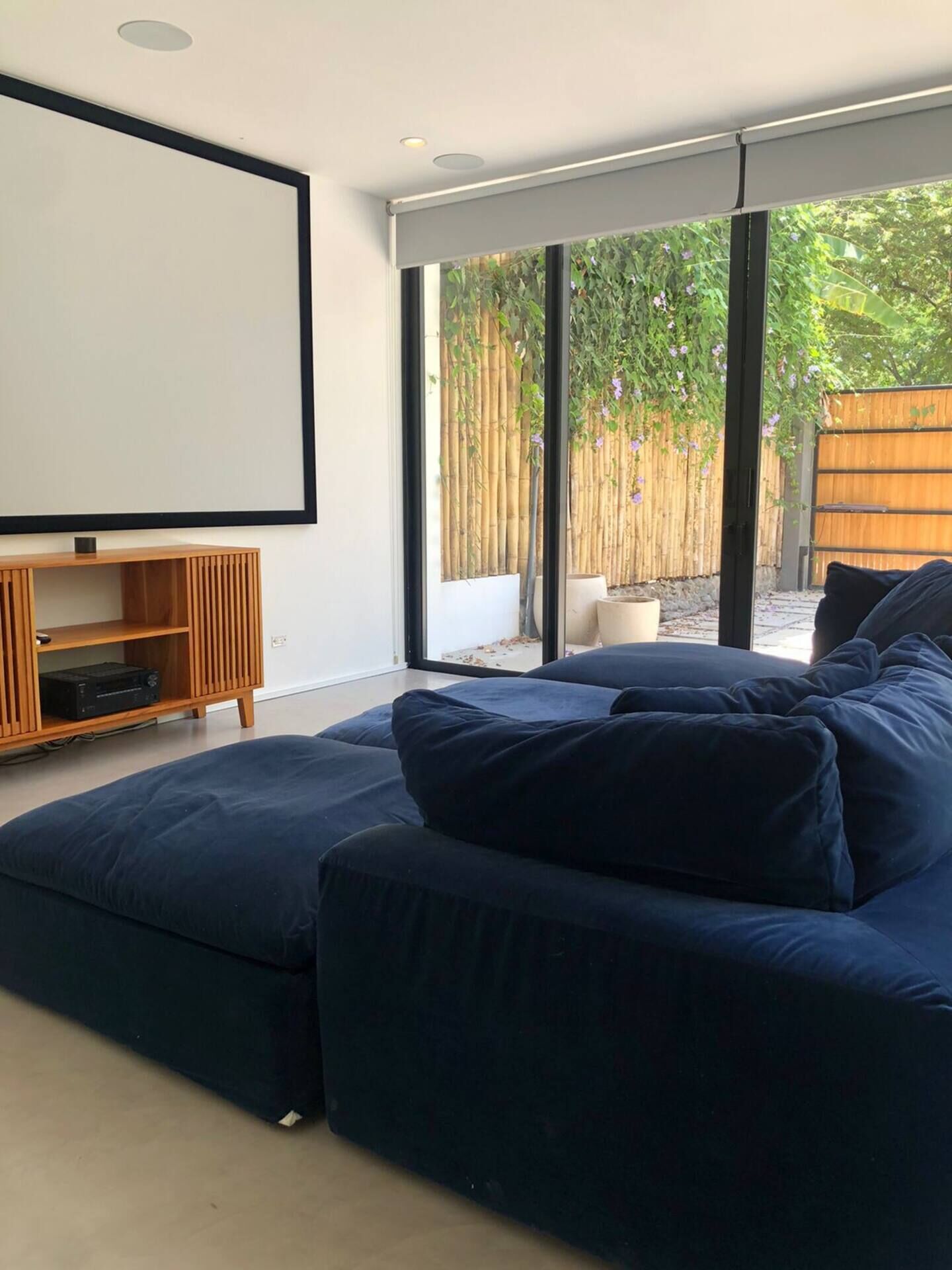 Ein modernes Wohnzimmer mit einem großen, dunkelblauen Modulsofa, einer Holzunterhaltungsecke und einem großen Fenster mit Blick auf einen Bambuszaun und viel Grün.