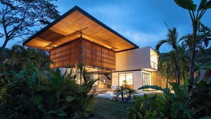Une maison moderne dotée d’une grande structure en bois en surplomb, entourée d’une végétation tropicale luxuriante.