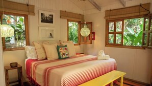 A bedroom with a bed, bedside table, bench, and large windows with views of greenery. - Mid Tide Bungalow - Walking distance to the Surf and Restaurants (Santa Teresa Beach)