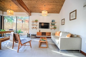 Relax in this spacious living room with a high ceiling, sofa, coffee table, and chair, featuring a flat-screen TV and large windows with a view of the outdoors.