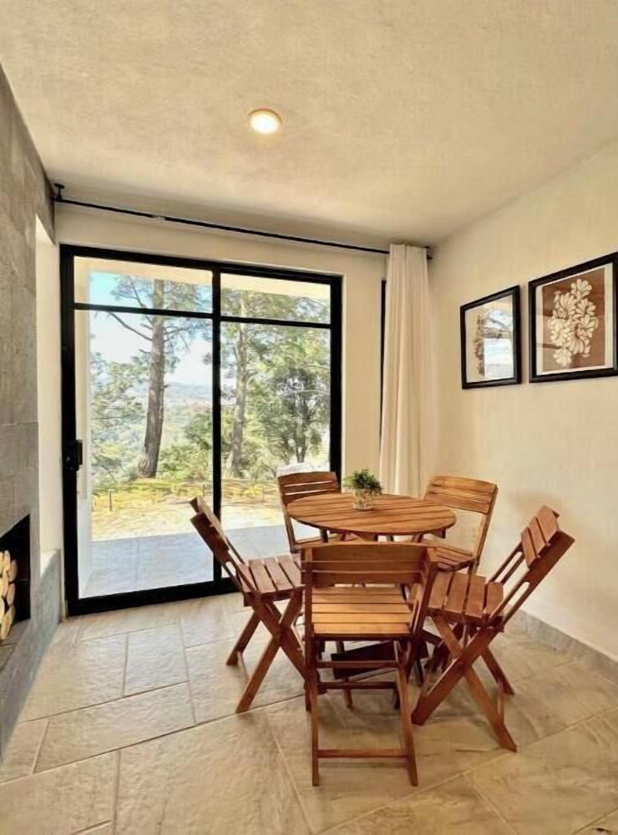 A dining area with a wooden table and chairs, a fireplace, and a large sliding glass door leading to a view of trees and hills.