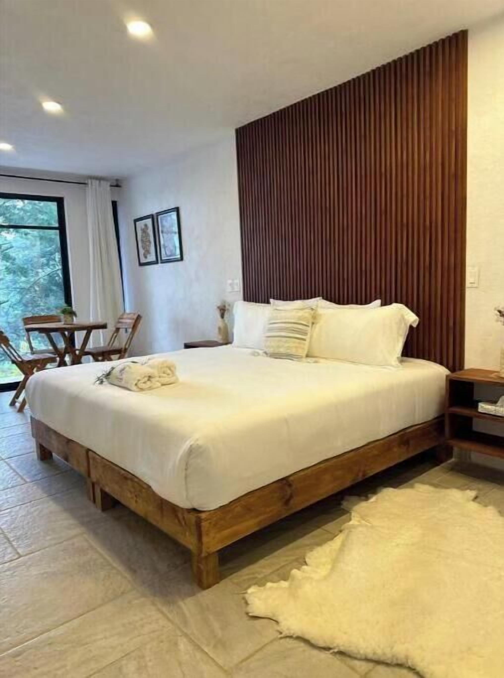 A bedroom with a wooden bed, a white rug, and a view of the outdoors through a window.
