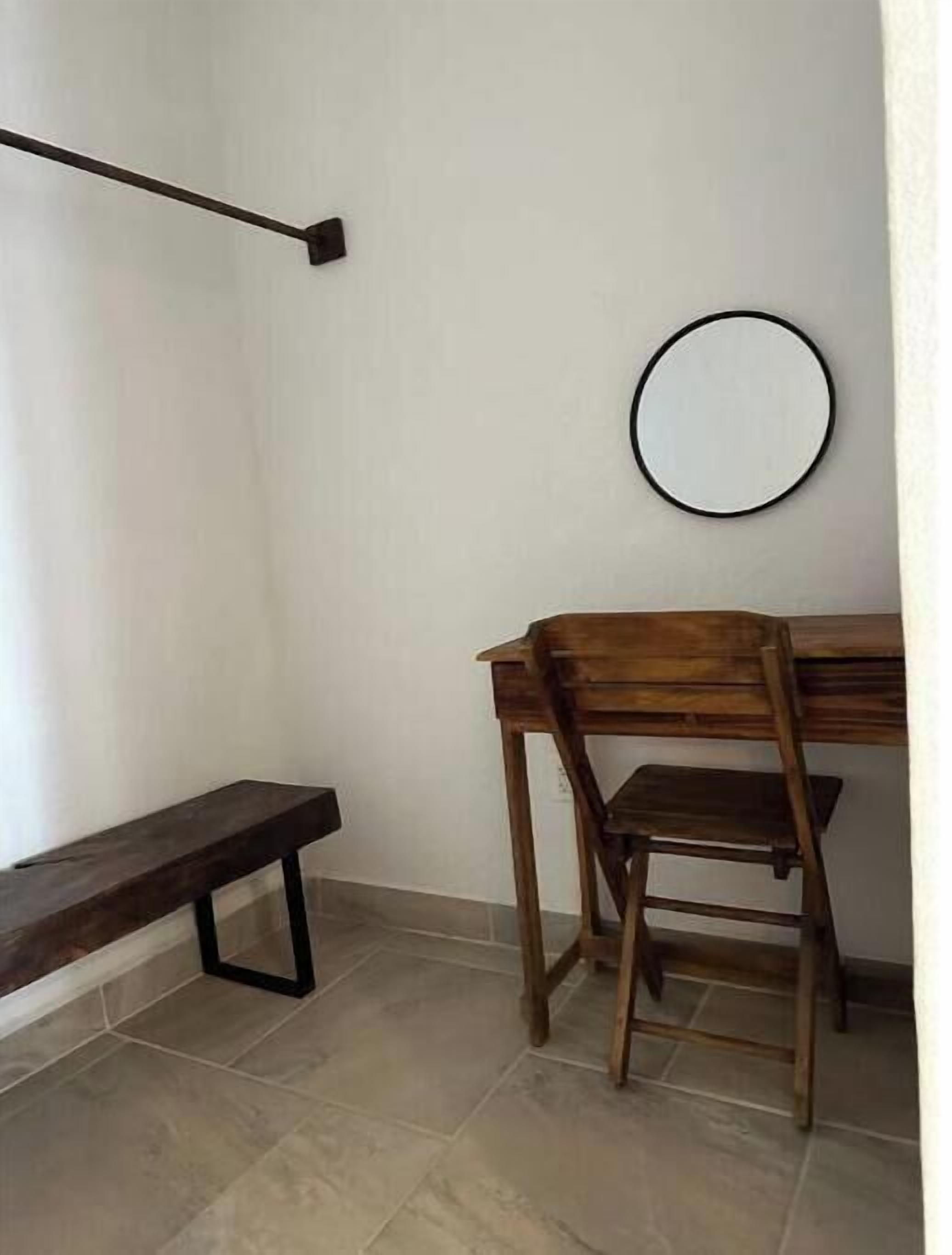 Une petite pièce simple, avec un banc en bois, un bureau avec une chaise et un miroir rond accroché au mur.
