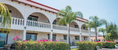 A hotel with a white facade, multiple balconies, and a gravel courtyard with benches and potted plants.