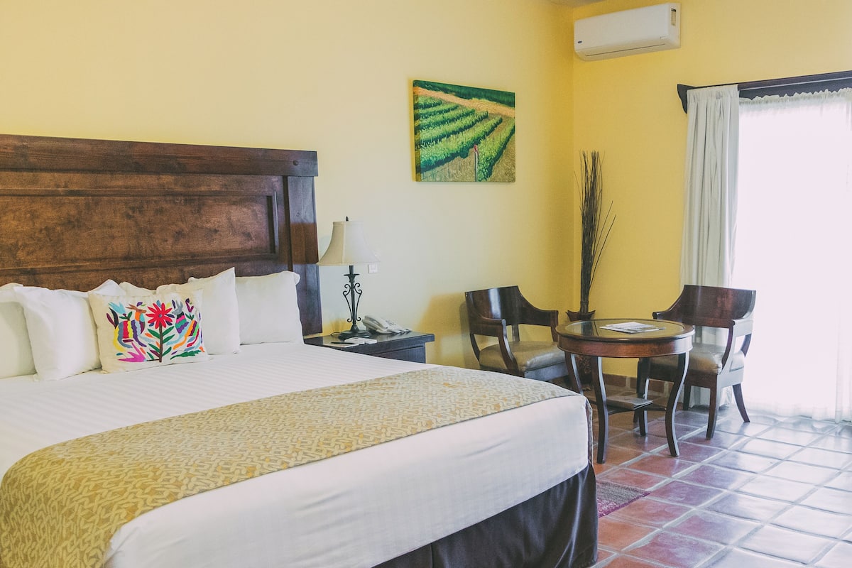A hotel room with a large bed, a small table with chairs, a painting on the wall, and an air conditioning unit.