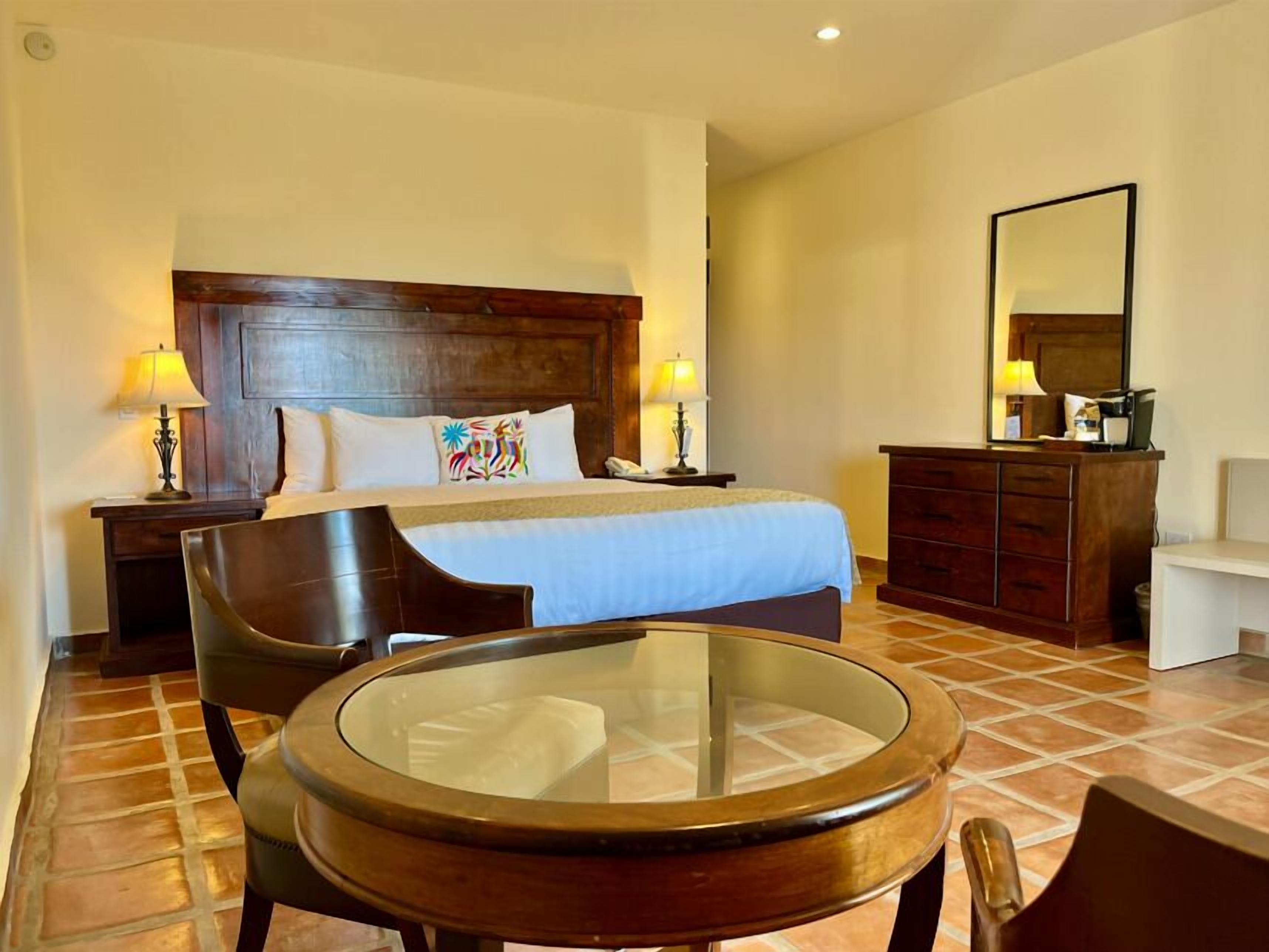 Habitación de hotel con una cama, dos mesitas de noche con lámparas, un aparador de madera, una mesa redonda de vidrio y una silla.
