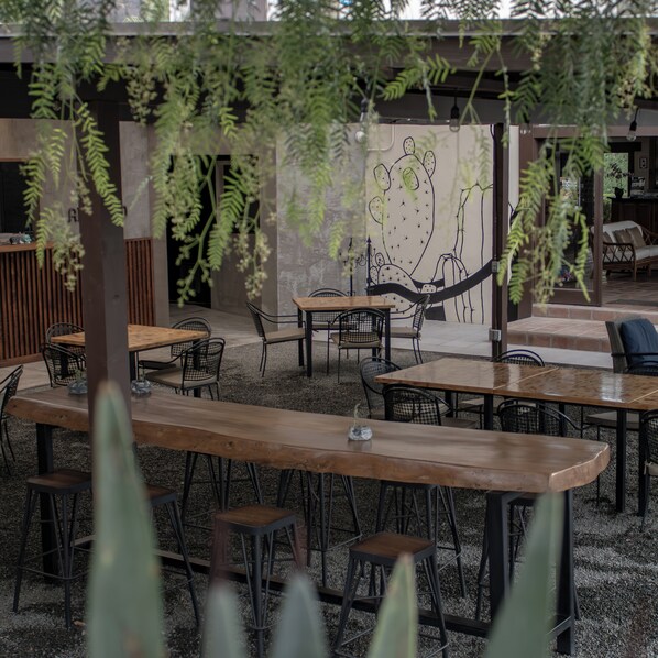 An outdoor seating area with tables and chairs, surrounded by greenery and a wall with a cactus mural. - Hotel Hacienda Guadalupe (Valle de Guadalupe)