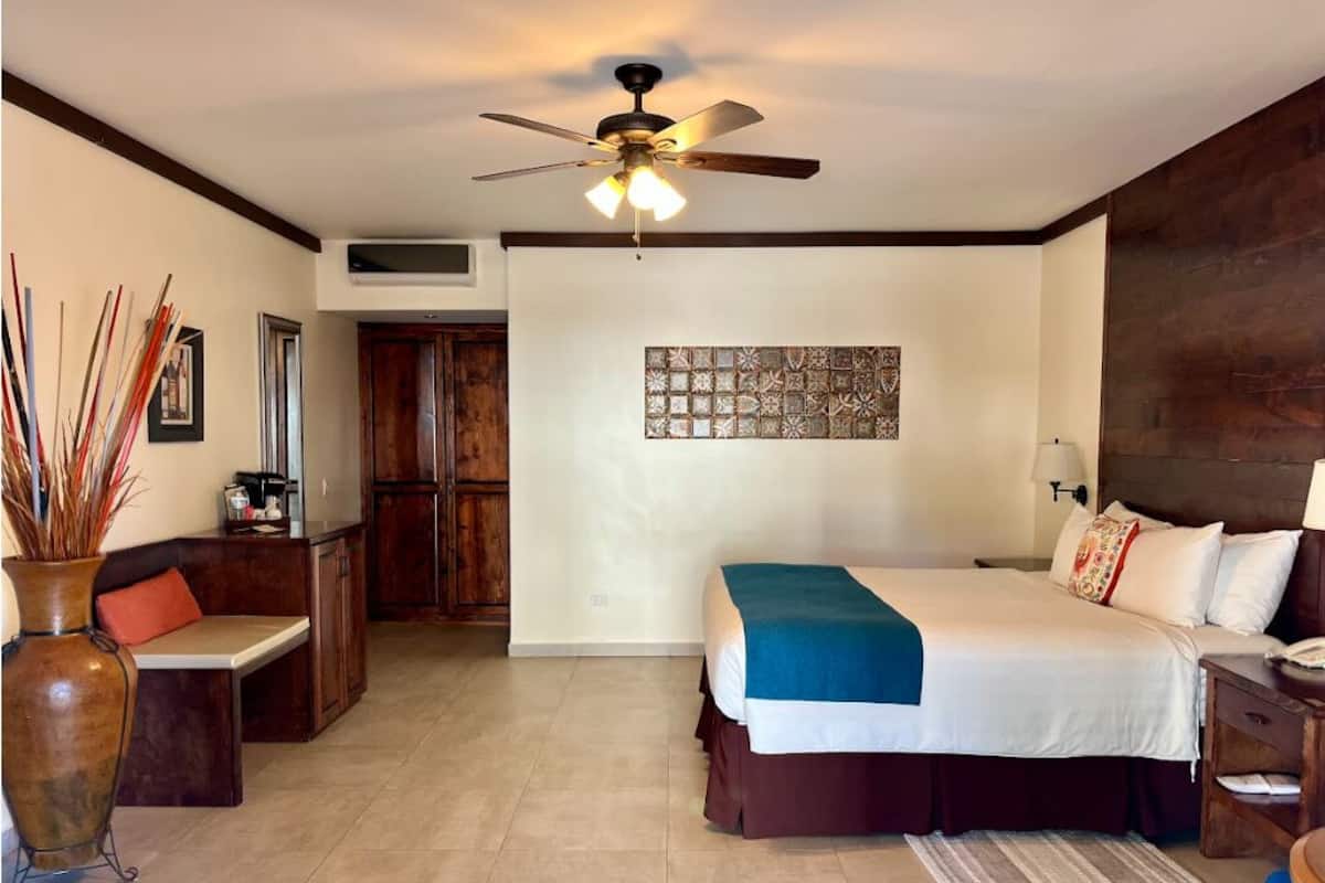 A bedroom with a bed, a ceiling fan, a wall-mounted air conditioner, a wooden cabinet, a decorative wall piece, and a vase with reeds.