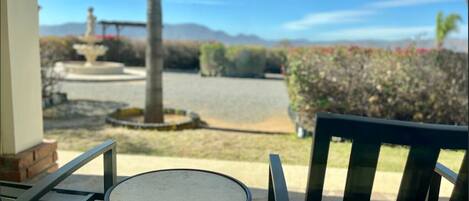 Un patio con un tavolo rotondo e sedie, una fontana e vista su un giardino curato con siepi e una montagna sullo sfondo.