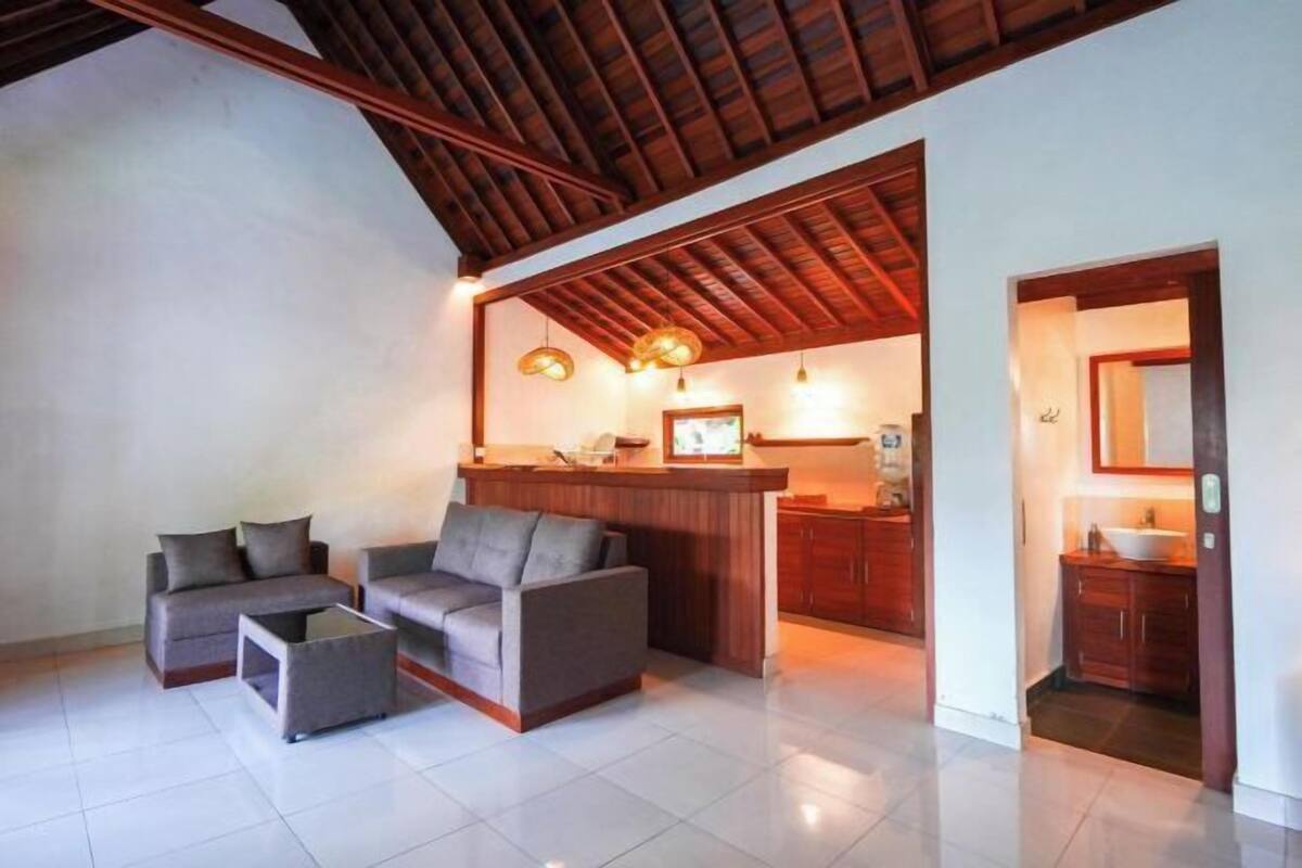 A spacious living area with a high ceiling and wooden beams, a kitchenette, and a bathroom with a mirror and sink.