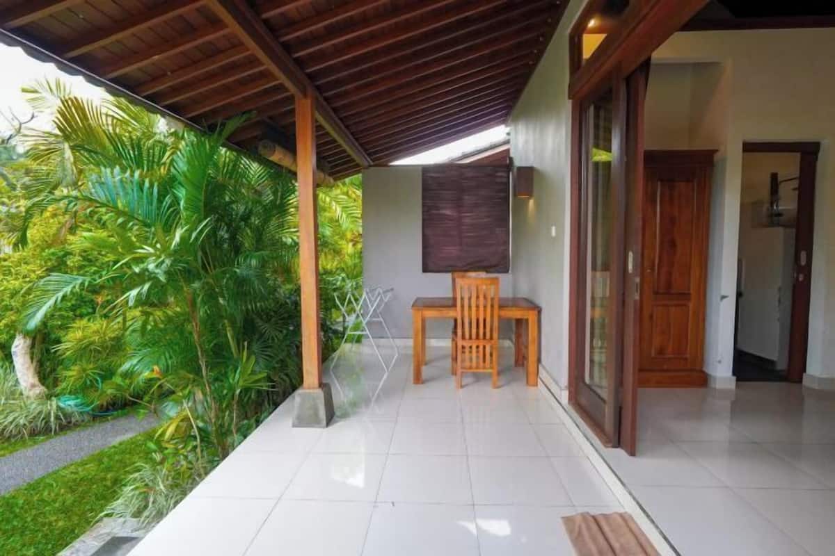 A covered patio with a wooden table and chair, surrounded by lush greenery.