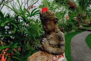 A statue of a seated figure with hands in a prayer position, surrounded by lush greenery and a curved pathway. - Green Coconut Cottage Ubud (Bali)