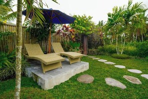 A garden with two wicker lounge chairs under a blue umbrella, surrounded by lush greenery and a bamboo fence. - Green Coconut Cottage Ubud (Bali)