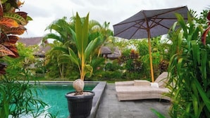 A pool area with a lounge chair and umbrella, surrounded by lush tropical plants. - Green Coconut Cottage Ubud (Bali)
