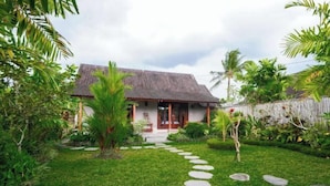 A house with a thatched roof, surrounded by lush greenery and a stone pathway leading to the entrance. - Green Coconut Cottage Ubud (Bali)