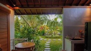 A compact kitchen with a small island, a dining area with a table and chairs, and a view of a lush garden with a stone path. - Green Coconut Cottage Ubud (Bali)