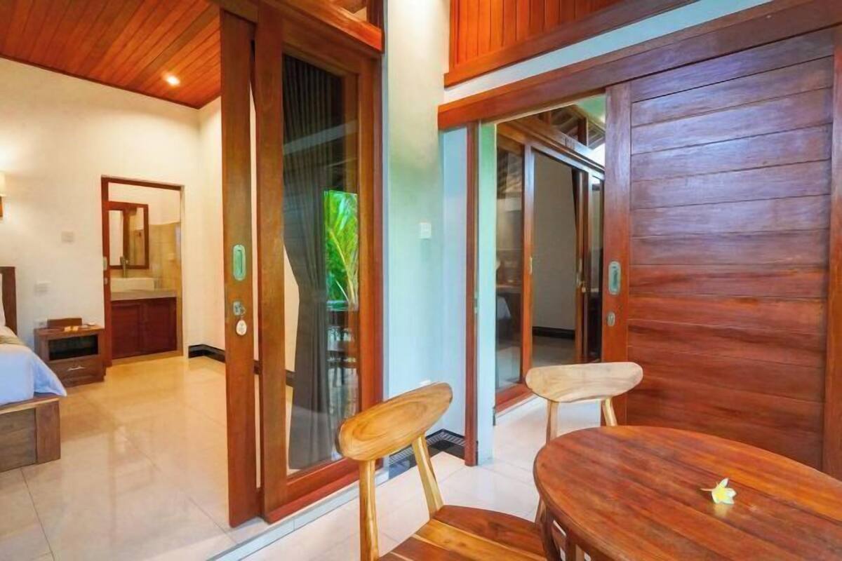 A room with a wooden ceiling, a bed, a desk, and a chair. There is a sliding glass door leading to another room.