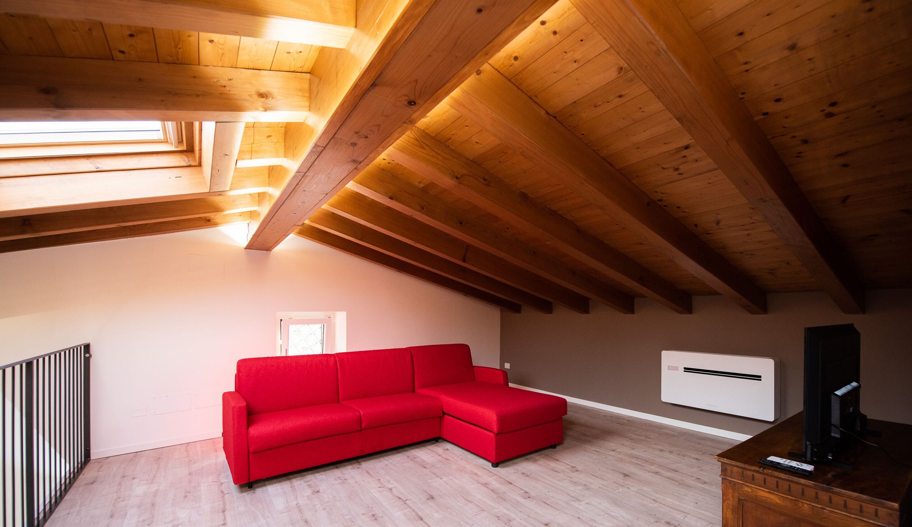 A room with a sloped wooden ceiling, a red sectional sofa, a white air conditioning unit, and a wooden TV stand.