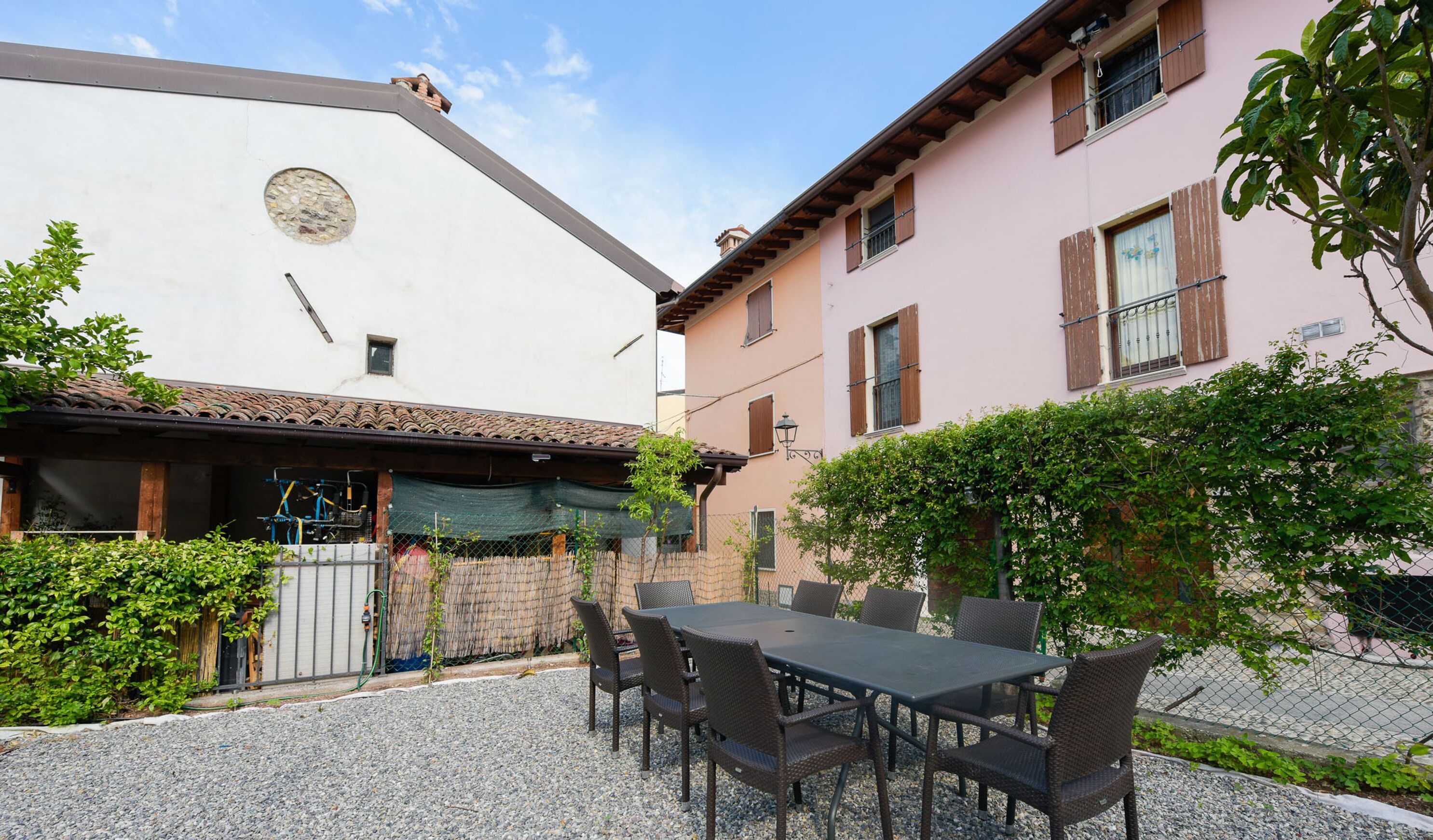 A courtyard with a table and chairs, surrounded by buildings with wooden shutters and a stone wall.