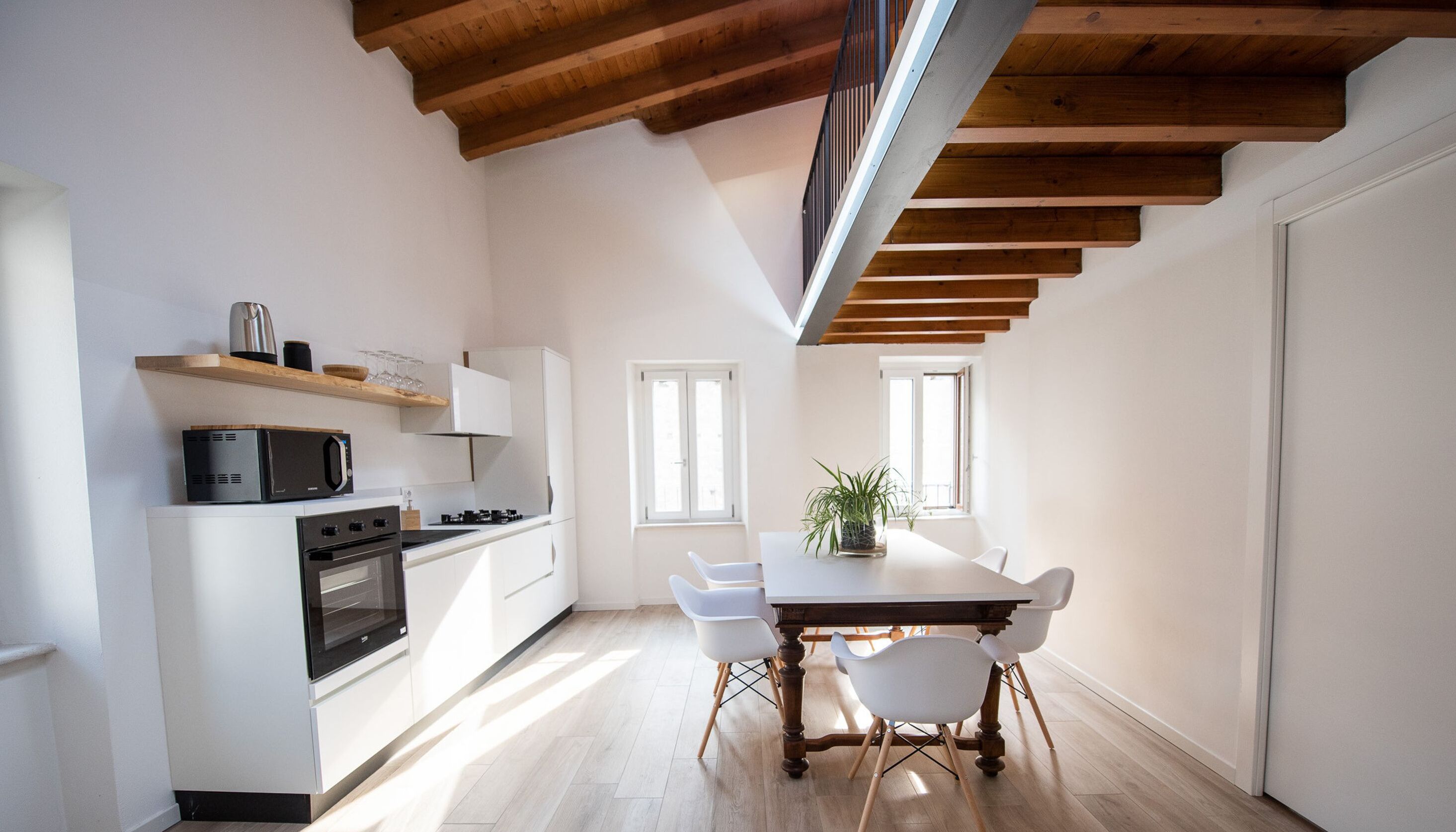 A modern kitchen with white cabinetry, a built-in oven, and a microwave. There is a dining table with chairs and a potted plant.