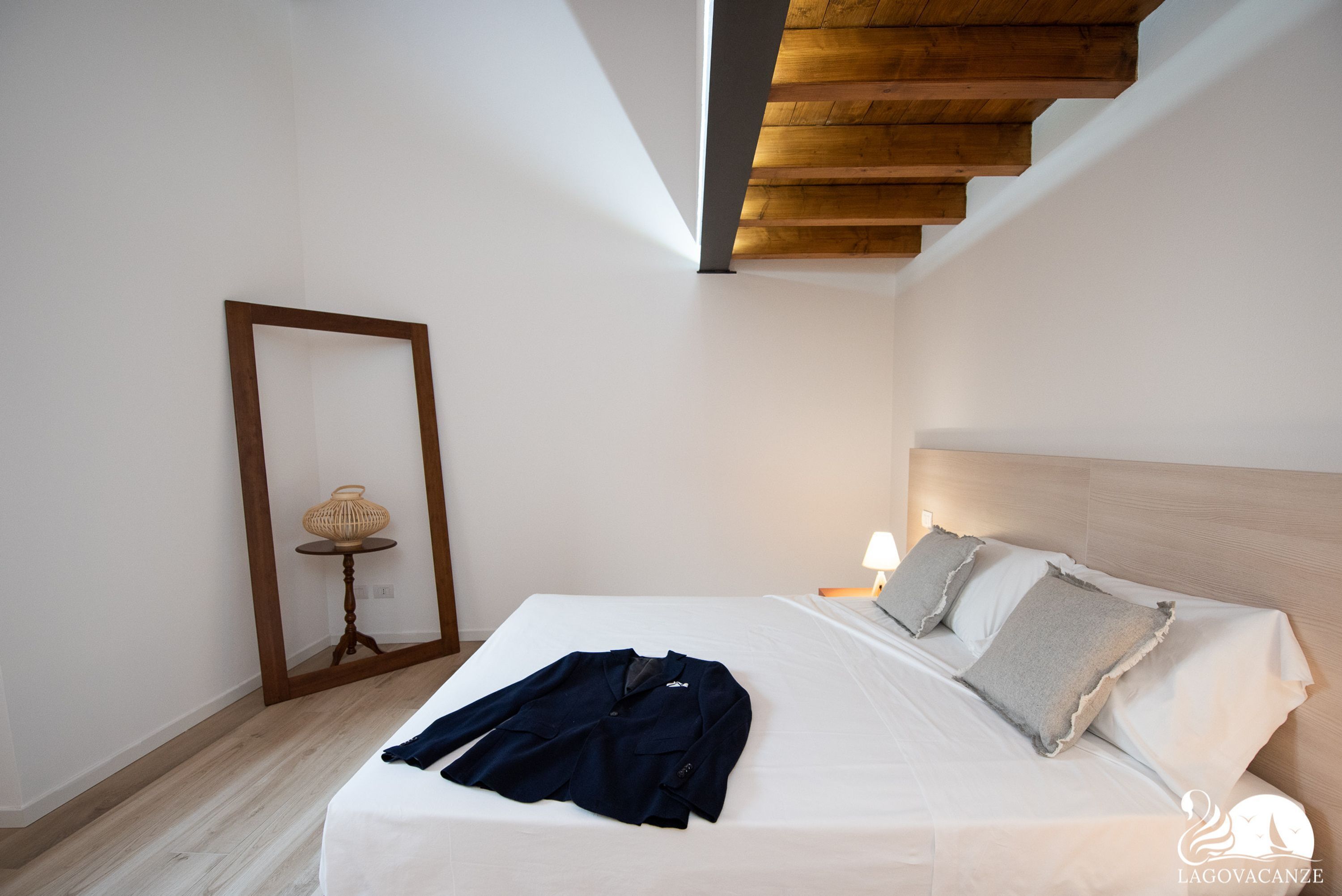 A bedroom with a wooden ceiling, a bed with white linens, a bedside table with a lamp, and a framed mirror on the wall.