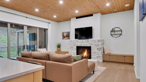 A modern living room with a stone fireplace, a flat-screen TV, and a large window overlooking a balcony. - Modern home with 2 master bedrooms, private hot tub, garage and mountain views (Silver Star Mountain)