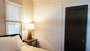 Un dormitorio con cama, mesita de noche con lámpara, ventana con persianas y una puerta.