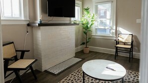 Una sala de estar con chimenea, televisor montado arriba, dos sillones, una mesita redonda, una planta en maceta y una ventana con persianas.