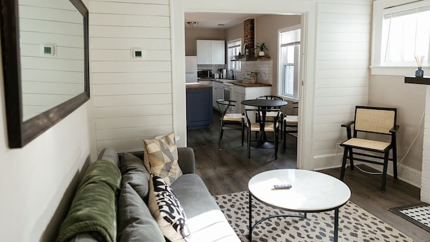 Una sala de estar acogedora con un sofá, una mesa de centro y una silla. Se ve una cocina al fondo con una zona de comedor.