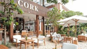 Un espacio al aire libre con mobiliario de madera y sombrillas, un letrero que dice "CORK" y un edificio de diseño moderno al fondo.
