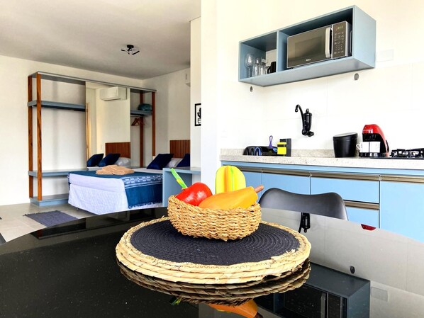 Une chambre d’hôtel compacte comprenant un lit, une kitchenette et un coin repas agrémenté d’un panier de fruits.