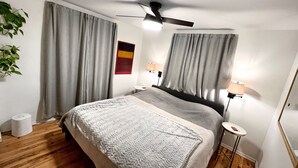 Un dormitorio con cama, cortinas, ventilador de techo, lámparas de noche, una mesita y una planta.