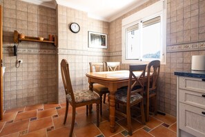 A dining area with a wooden table and chairs, a clock, a window with a view, and a tiled backsplash.