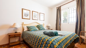 Un dormitorio con una cama, dos mesitas de noche, un ventanal con cortinas y tres cuadros enmarcados en la pared.