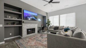 A modern living room with a fireplace, a flat-screen TV, a ceiling fan, and a large window with blinds. - Charming 4-bedroom house with private pool and spa in brilliant Indio (Indio)