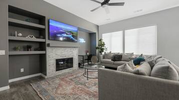 A modern living room with a fireplace, a flat-screen TV, a ceiling fan, and a large window with blinds.