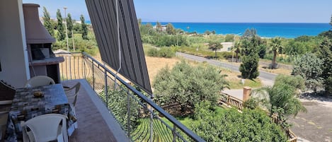 A balcony with a table and chairs, overlooking a coastal landscape with trees and a road.