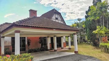 A single-story building with a covered entrance, a chimney, and a garden with tropical plants.