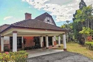 Un edificio de una sola planta con un porche cubierto, una chimenea y un jardín con plantas tropicales.