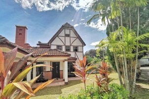 Una casa con techo a dos aguas, una chimenea y un jardín bien cuidado con plantas tropicales.