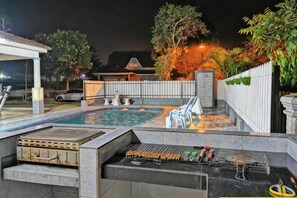 Un área de piscina al aire libre con una parrilla, sillones de descanso y una zona de estar cubierta.
