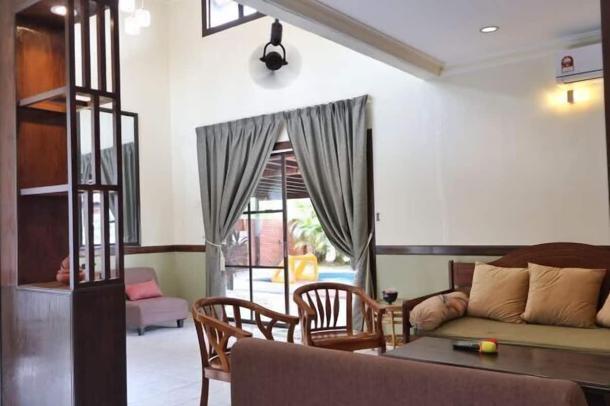 A room with a sofa, two wooden chairs, a small table, and a view of a pool through a window with curtains.