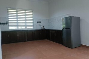 Una cocina con armarios oscuros, un refrigerador, una ventana con persianas y piso de baldosas.