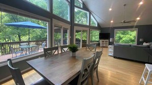 A spacious living area with a large wooden table, multiple chairs, a sofa, and a TV. There are large windows offering views of trees. - River Relaxation - Calm Riverfront Home in Winchester Kentucky (Winchester)