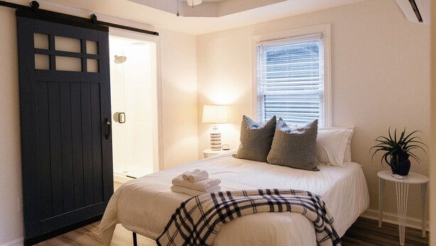 A bedroom with a bed, a bedside table with a lamp, a window with blinds, a ceiling fan, and a dark sliding door. - Big Blue - Stylish Studio in Downtown Lexington (Lexington)